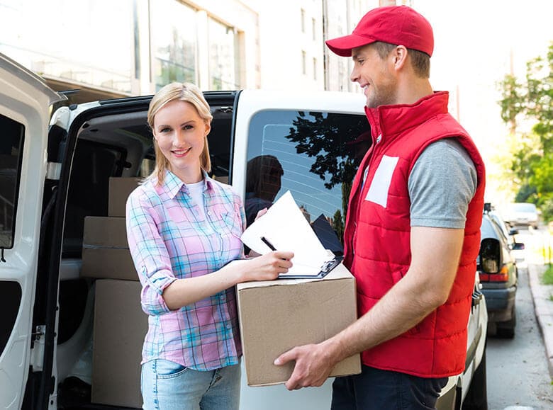 The image depicts the rear section of a white Hire Man and Van removal van parked on a paved driveway in a residential area, with the vehicle's side door closed. The van's exterior appears clean and in good condition, with subtle reflections of surrounding trees and nearby buildings visible on its surface. To the right of the van, there is a pile of mixed waste materials, including several black rubble sacks and a collection of loose items such as wooden planks, cardboard boxes, and plastic containers, all stacked against the side of the vehicle. The ground is a grey concrete paving surface, and a small section of a garden or yard with greenery is visible in the background. Overhead, the sky is overcast, emitting diffuse natural light that softens shadows and highlights the textures of the waste and the van's exterior. The scene captures a typical rubbish removal setup, illustrating a local move or clearance within the Kildary area, as managed by Hire Man and Van's professional service team specializing in removals and waste disposal solutions in the IV18 postcode.